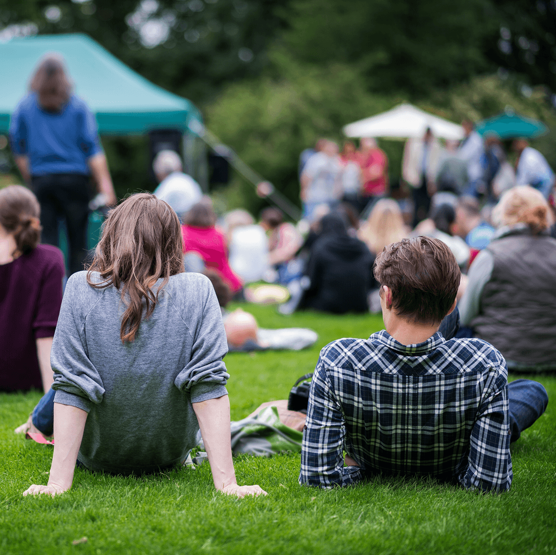 People sitting on grass at an outdoor community event with tents and crowds in the background