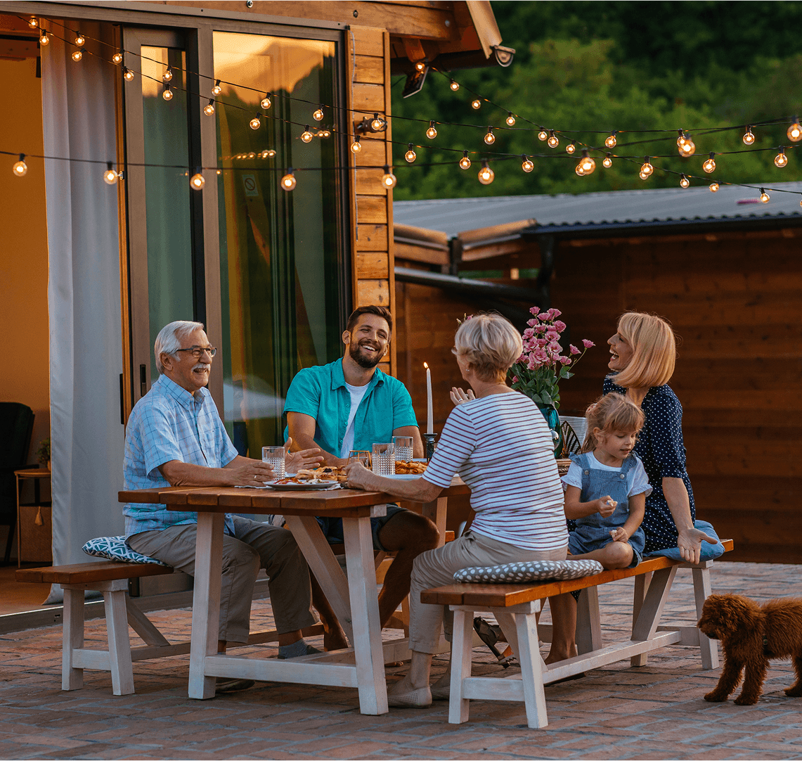 Smiling family enjoying dinner together on a patio with string lights and a small dog nearby