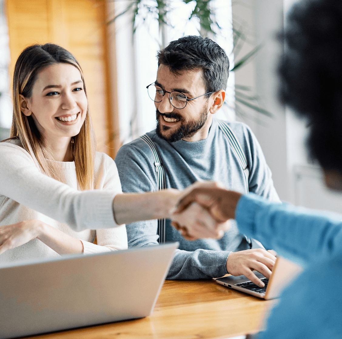Smiling woman shaking hands across a table while sitting with a man using a laptop